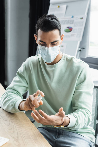 Μουσουλμάνος επιχειρηματίας με ιατρική μάσκα που χρησιμοποιεί απολυμαντικό χεριών στο γραφείο  - Φωτογραφία, εικόνα