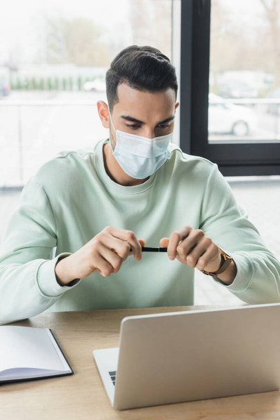 Arabian businessman in medical mask holding pen near laptop in office  - Φωτογραφία, εικόνα