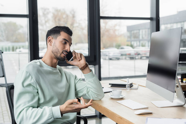 Hombre de negocios musulmán hablando en el teléfono inteligente cerca de la computadora y el café en la oficina  - Foto, imagen