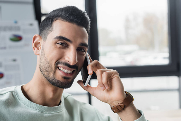 Улыбающийся мусульманский бизнесмен разговаривает на смартфоне и смотрит в камеру в офисе  - Фото, изображение