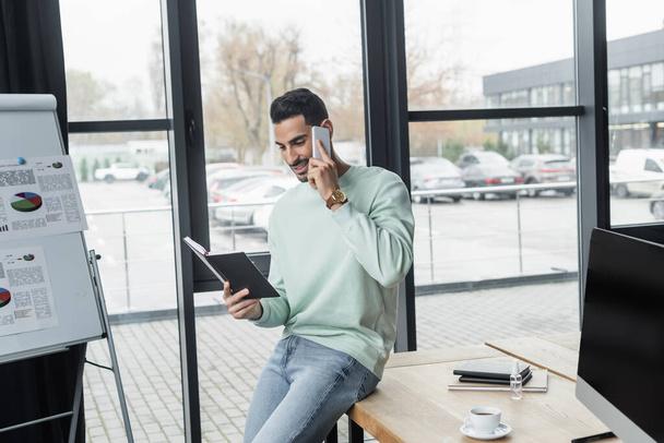 Pozitivní arabský podnikatel při pohledu na notebook, zatímco mluví na smartphone v kanceláři  - Fotografie, Obrázek