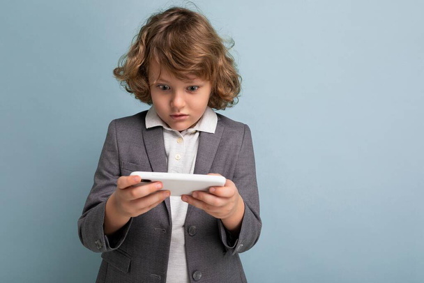 Zdjęcie przystojnego emocjonalnego chłopca z kręconymi włosami, noszącego szary garnitur i korzystającego z telefonu odizolowanego na niebieskim tle, patrzącego na smartfona grającego w gry - Zdjęcie, obraz