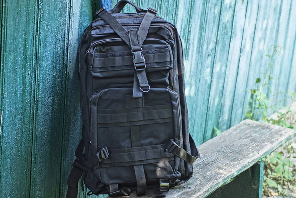 one black tactical backpack stands on a gray wooden bench against a green wall on the street - Photo, Image