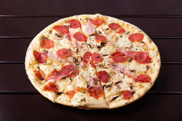 A whole rustic pizza with mozzarella, brisket, mushrooms and salami is served on a paper plate on a wooden table - Photo, Image
