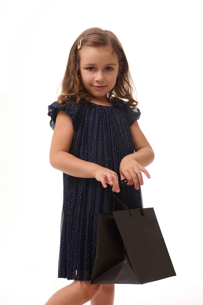 Confident portrait isolated on white background with copy space of pretty baby girl looking at camera with black shopping packet in her hands,dressed in evening dark blue dress. Black Friday concept - Fotografie, Obrázek