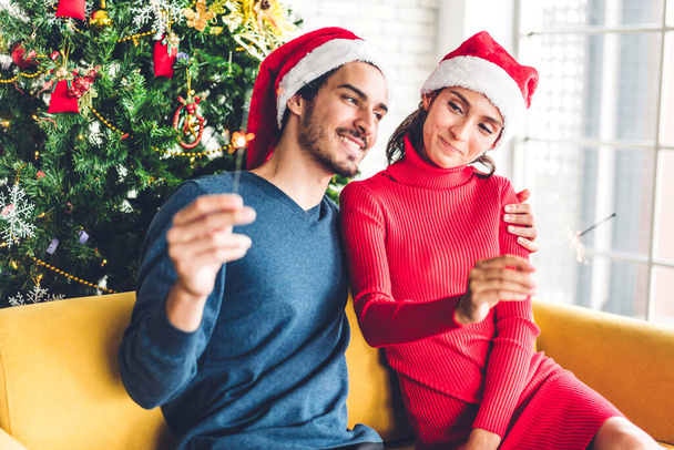 Romantic sweet couple in santa hats having fun decorating christmas tree and smiling while celebrating new year eve and enjoying spending time together in christmas time at home - Photo, Image