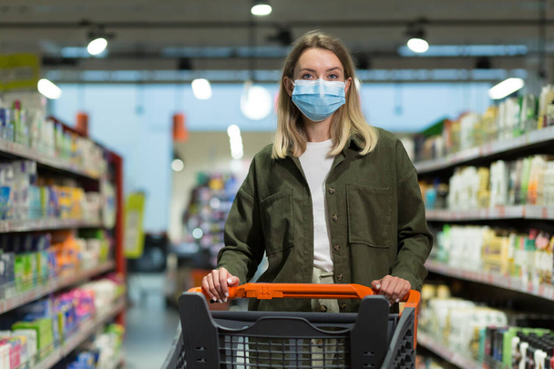woman wearing medical face mask push shopping cart in supermarket. Young girl choosing, looking grocery things to buy. Girl walks through supermarket or store. joyful Pretty female walking in mall - 写真・画像