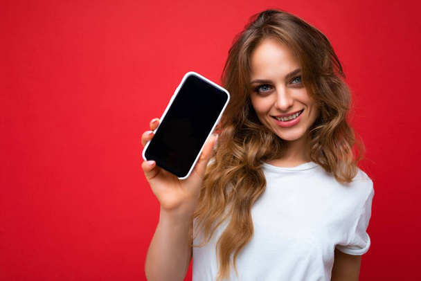 Kuva kaunis hymyilevä nuori blondi nainen hyvännäköinen yllään valkoinen t-paita seisoo eristetty punaisella taustalla kopio tilaa tilalla puhelin osoittaa älypuhelin kädessä tyhjä näyttö - Valokuva, kuva