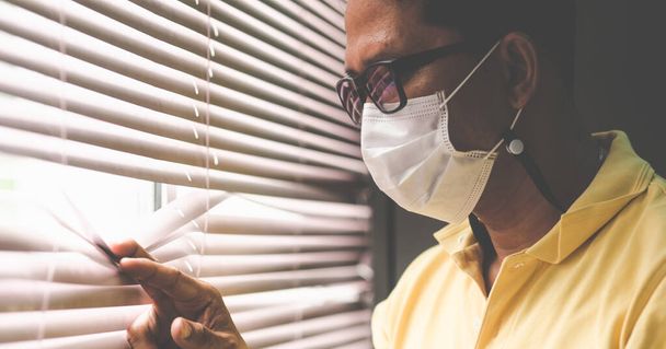 Een man met een medisch masker die uit het raam kijkt, werkt thuis vanwege de virusuitbraak, nieuw normaal.. - Foto, afbeelding