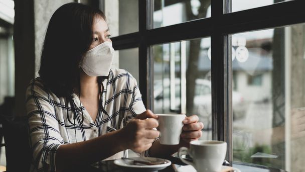 Eine Frau mit medizinischer Maske sitzt in einem Café und blickt mit traurigen Augen aus dem Fenster. Sie hat genug vom Ausbruch des Virus und will ein normales Leben führen.. - Foto, Bild