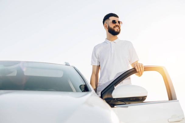 Stylish young handsome man in sunglasses posing near a car in the city center, Handsome man near a modern white car on a city street - Fotografie, Obrázek