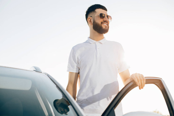 Stylish young handsome man in sunglasses posing near a car in the city center, Handsome man near a modern white car on a city street - Foto, Bild