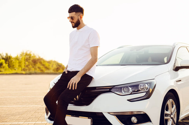 Handsome young man with a beard, stands next to the car, Handsome man with a white car, Photo of a sexy young man with a classic car outdoors on a sunny day - Fotó, kép