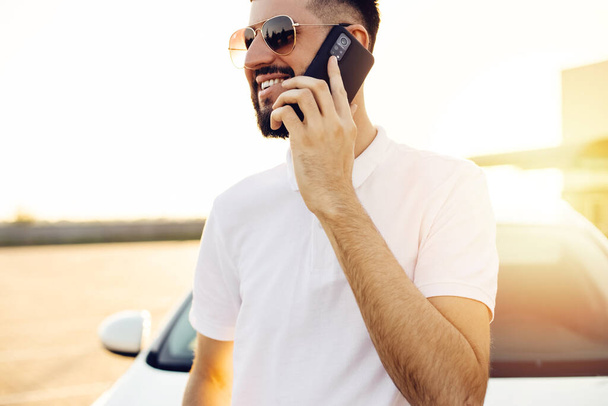 Handsome young business man talking on a mobile phone in the city near a white car, a man in glasses posing near a car in the city outdoors - Foto, Bild
