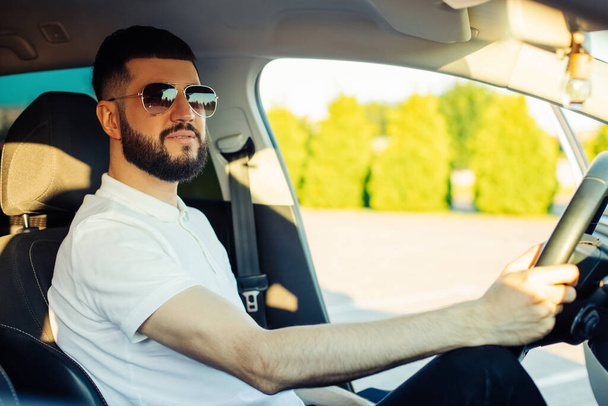 Happy young man, wearing sunglasses, sits in the driver's seat and smiles, Taxi driver listens to music in the car and drives - Fotografie, Obrázek