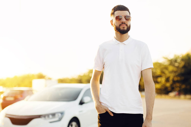 Handsome young man, businessman in sunglasses and white shirt, stands near his sports car, man near his car, outdoors - Foto, afbeelding