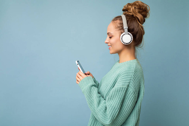 Zdjęcie pięknej, radosnej, uśmiechniętej młodej kobiety w stylowych, luźnych ciuchach odizolowanych nad ścianą tła, trzymającej i używającej telefonu komórkowego w białych słuchawkach bluetooth słuchających muzyki i - Zdjęcie, obraz