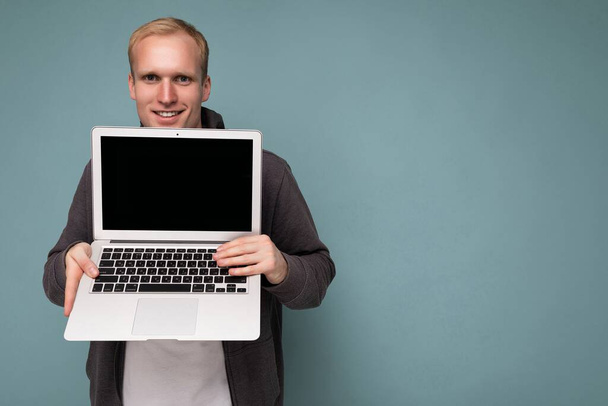 Retrato de primer plano del guapo hombre rubio sonriente sosteniendo computadora portátil con pantalla de monitor vacía con maqueta y espacio de copia mirando cámara aislada sobre fondo azul - Foto, Imagen