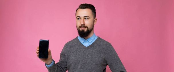 Panoramatické foto pohledný šťastný cool mladé brunetky neoholený muž s vousy na sobě stylový šedý svetr a modrá košile stojící izolované přes růžové pozadí zdi drží smartphone a ukazuje - Fotografie, Obrázek