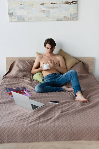 shirtless transgender young man in jeans holding cup of coffee while watching movie on laptop in bedroom  - Foto, Bild