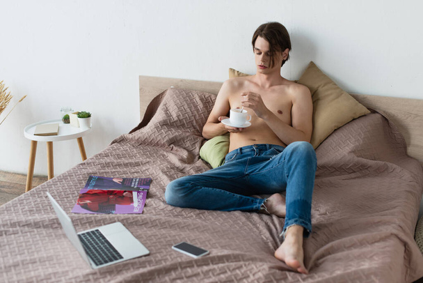 barefoot transgender young man in jeans holding cup of coffee near gadgets on bed - Foto, afbeelding
