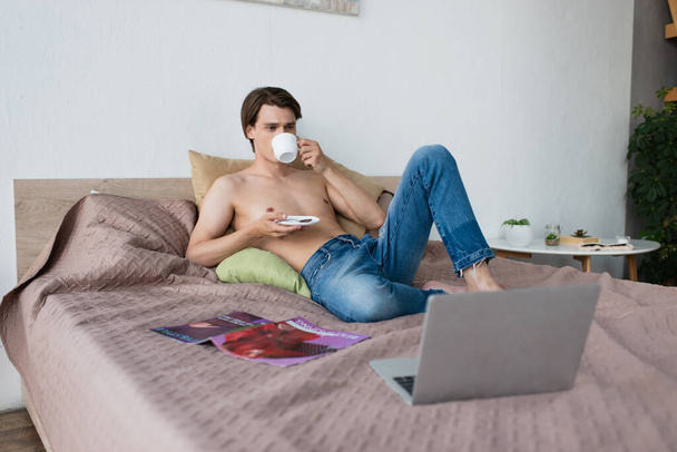 shirtless transgender young man in jeans holding cup and drinking coffee near laptop on bed - 写真・画像