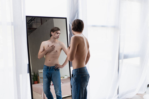 félmeztelen transznemű fiatalember farmerban, kéz a csípőn áll, miközben tükörbe néz  - Fotó, kép