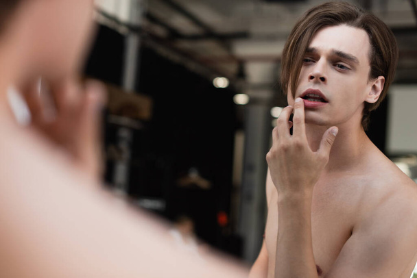 shirtless transgender young man touching lip and looking at mirror  - Foto, Bild