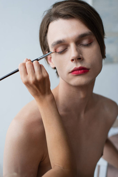 makeup artist applying eye shadow with cosmetic brush on transgender man  - Foto, Imagen