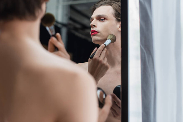 young transgender man applying face powder on neck near mirror  - 写真・画像