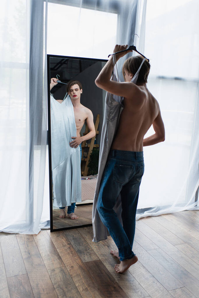 teljes hossza fiatal transznemű férfi smink gazdaság vállfa csúszás ruha tükör közelében  - Fotó, kép