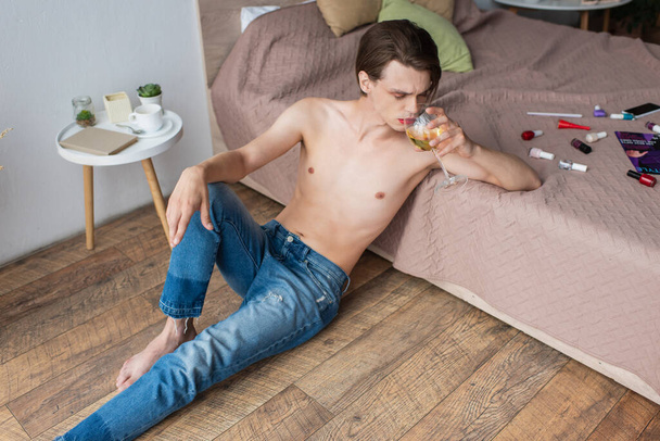 fiatal transznemű férfi bort iszik, miközben ül az ágy mellett smink termékek - Fotó, kép