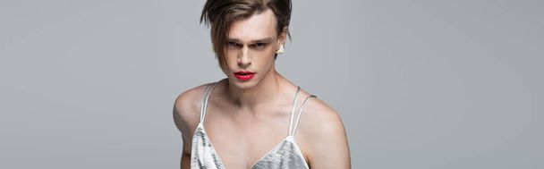 νεαρός τρανσέξουαλ άνδρας με μακιγιάζ και σκουλαρίκι κοιτάζοντας κάμερα απομονωμένη σε γκρι, banner - Φωτογραφία, εικόνα