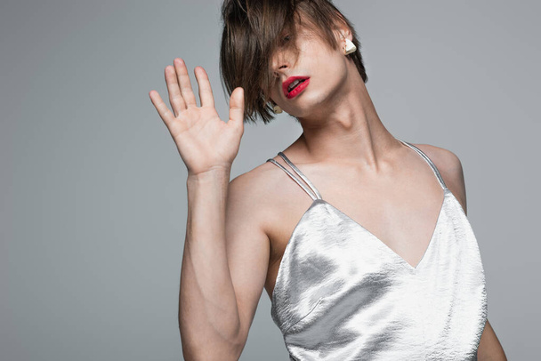 young transgender man with red lips and earring posing in slip dress isolated on gray - Фото, изображение