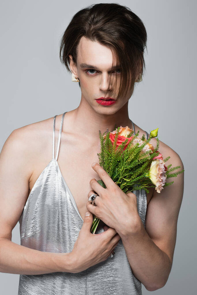 young transgender man in slip dress holding flowers isolated on gray - Zdjęcie, obraz
