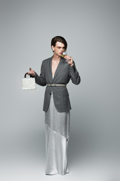 πλήρες μήκος του τρανσέξουαλ άνθρωπος σε ολίσθηση φόρεμα και σακάκι πόσιμο κρασί, ενώ κρατώντας τσάντα σε γκρι - Φωτογραφία, εικόνα