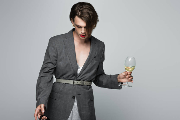 young transgender man in blazer holding glass of wine isolated on grey - Photo, Image