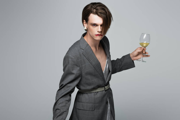 młody transgenderowy mężczyzna w marynarce gryzie wargę trzymając kieliszek wina odizolowanego na szarości - Zdjęcie, obraz
