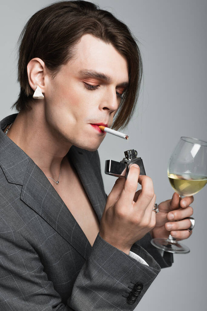 молодой трансгендер с бокалом вина и зажигалки во время курения изолированы на сером - Фото, изображение