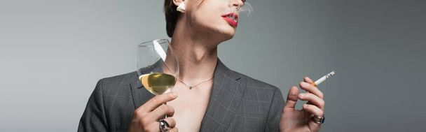 oříznutý pohled na mladého transsexuála v saku a náušnici se sklenicí vína při kouření na šedé, transparentní - Fotografie, Obrázek