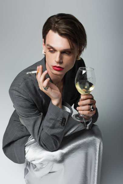 young transsexual man in blazer and slip dress holding glass of wine while smoking cigarette on grey - Photo, image