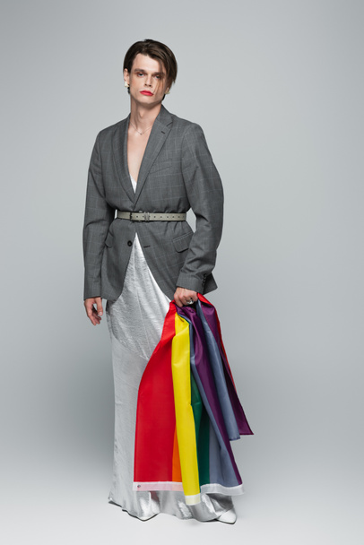 full length of young transsexual man in blazer and slip dress holding lgbt flag on grey - Fotó, kép