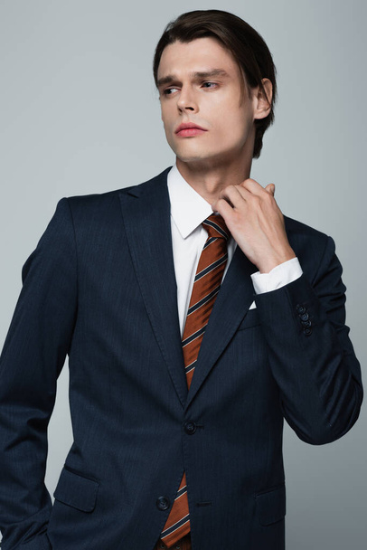 young androgynous man in blazer and tie adjusting collar isolated on grey - Fotó, kép