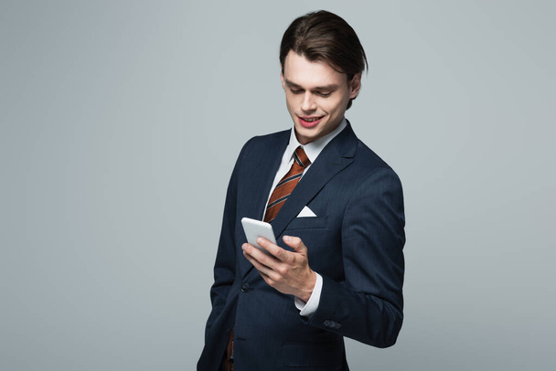 cheerful young businessman in suit using smartphone isolated on grey - Foto, Imagem