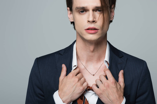 young transsexual businessman in suit adjusting shirt and showing necklace isolated on grey - Foto, afbeelding