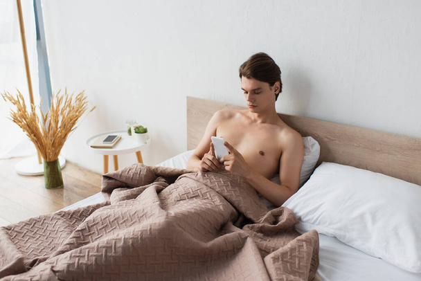 homme transgenre torse nu en utilisant un smartphone au lit - Photo, image