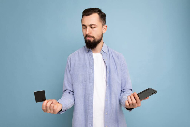 Fotografie dobře vypadající atraktivní brunet neoholený mladý muž na sobě ležérní modrá košile a bílé tričko izolované přes modré pozadí zdi drží kreditní kartu a pomocí mobilního telefonu platby - Fotografie, Obrázek