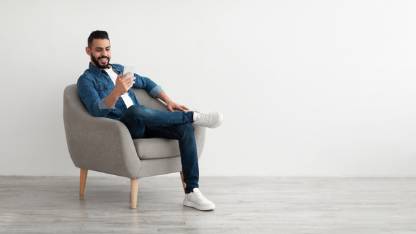 Cheerful young Arab man having online business meeting on smartphone, sitting in cozy armchair near white wall - Foto, Bild