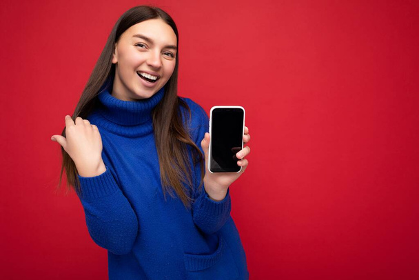 Portré mosolygós gyönyörű boldog fiatal barna nő visel alkalmi kék pulóver elszigetelt piros háttér üres hely tartja a kezében mobiltelefon és mutatja okostelefon üres képernyőn - Fotó, kép