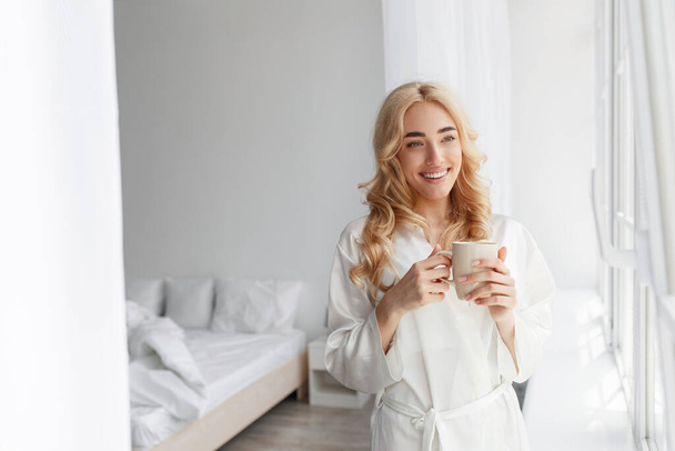 Iloinen nuori söpö eurooppalainen blondi nainen kylpytakissa nauttia hyvää huomenta, rauhaa, hiljaisuutta ja juoda herkullista tuoretta kahvia. - Valokuva, kuva